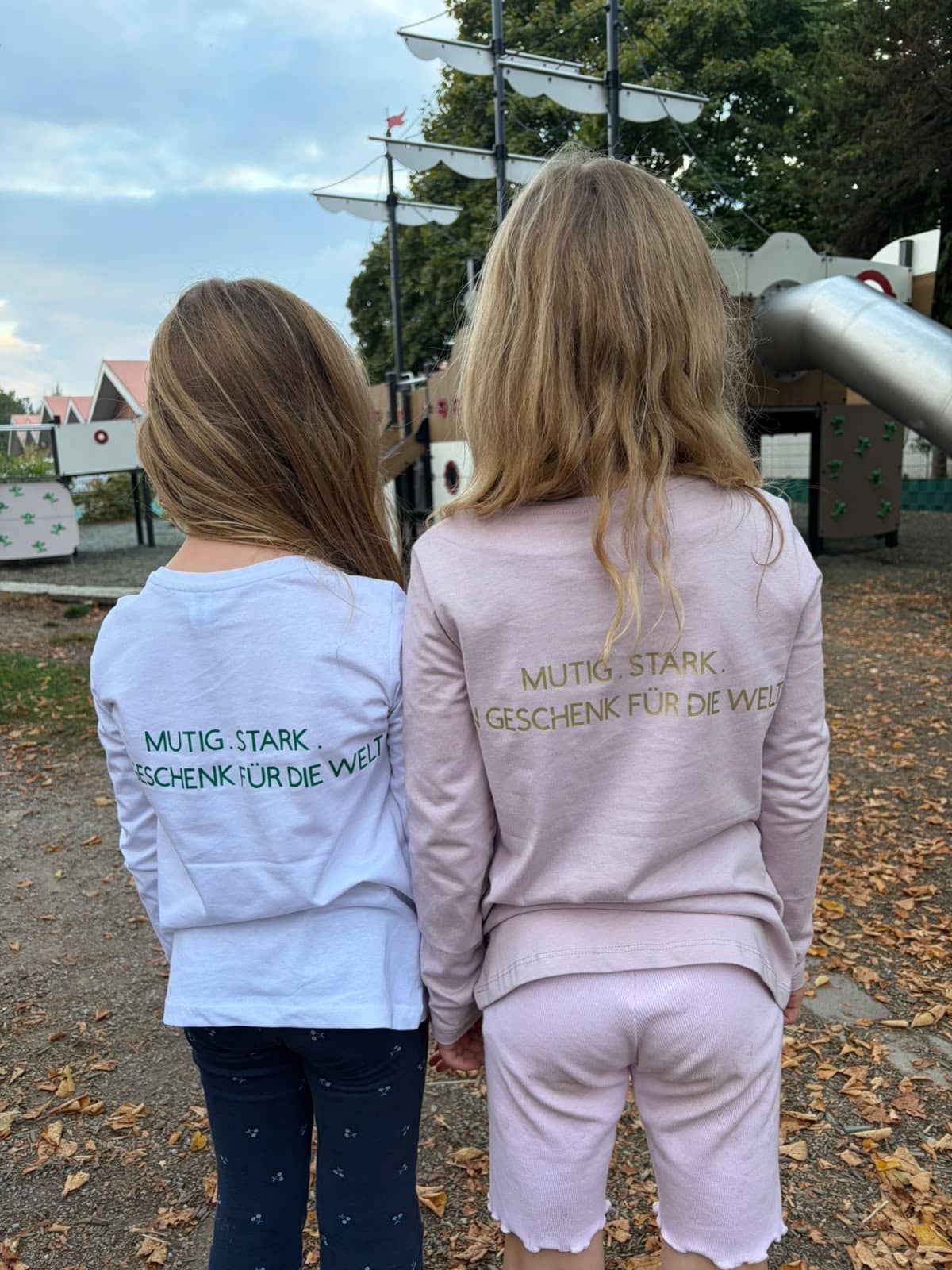 Zwei Kinder von hinten auf dem Spielplatz, auf ihren Shirts steht: Mutig. Stark. Ein Geschenk für die Welt.